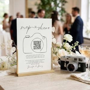 May include: A white rectangular sign with a gold metal stand featuring the black text "snap & share" above a line-art camera icon containing a QR code. Below the icon, text reads "YOUR PRESENCE MEANS THE WORLD TO US" and "Jennifer & Henry".