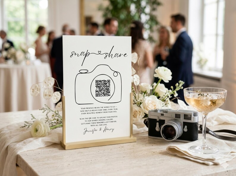 May include: A white rectangular sign with a gold metal stand featuring the black text "snap & share" above a line-art camera icon containing a QR code. Below the icon, text reads "YOUR PRESENCE MEANS THE WORLD TO US" and "Jennifer & Henry".