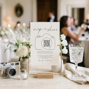 May include: A white rectangular sign with black text and a line-art camera icon featuring a QR code. The sign reads "snap & share" and includes instructions for guests to upload photos. It is held in a gold metal stand on a wooden base.