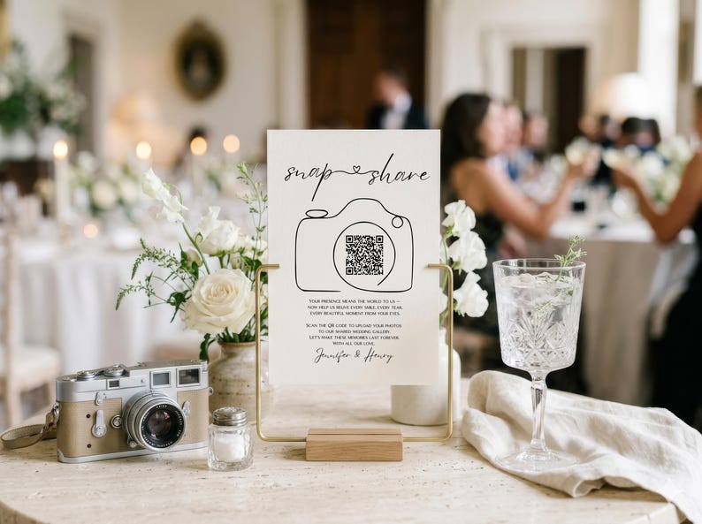 May include: A white rectangular sign with black text and a line-art camera icon featuring a QR code. The sign reads "snap & share" and includes instructions for guests to upload photos. It is held in a gold metal stand on a wooden base.