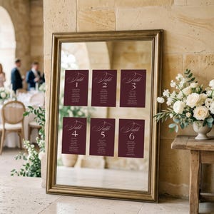 Puede incluir: Un espejo rectangular grande con marco dorado que muestra seis planos de mesa de color burdeos etiquetados del Table 1 al Table 6 en fuente cursiva blanca. El espejo refleja una recepción de boda con mesas, sillas e invitados.
