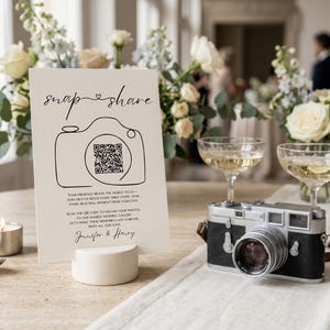 May include: A white rectangular wedding sign featuring the black text "snap & share" above a line-art camera illustration containing a QR code. Below, the text reads "Your presence means the world to us" and "Jennifer & Henry" in a script font.
