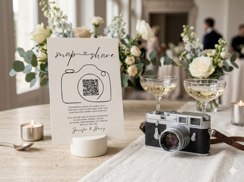 May include: A white rectangular wedding sign featuring the black text "snap & share" above a line-art camera illustration containing a QR code. Below, the text reads "Your presence means the world to us" and "Jennifer & Henry" in a script font.