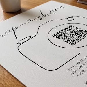 May include: A white sign featuring a black line drawing of a camera with a QR code in the center of the lens. The text "snap & share" is written in a cursive script above the camera, with additional text visible at the bottom right corner.