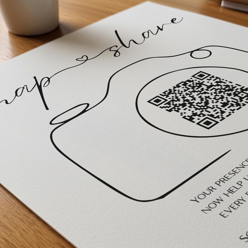 May include: A white sign featuring a black line drawing of a camera with a QR code in the center of the lens. The text "snap & share" is written in a cursive script above the camera, with additional text visible at the bottom right corner.