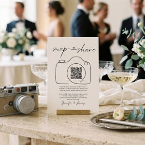 May include: A rectangular white wedding sign featuring the text "snap & share" above a line-art camera icon containing a QR code. Below the icon, the text reads "Your presence means the world to us - now help us relive every smile, every tear, every beautiful moment from your eyes. Scan the QR code to upload your photos to our shared wedding gallery. Let's make these memories last forever. With all our love, Jennifer & Henry."