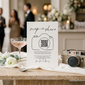 May include: A white rectangular wedding sign with black text reading snap share, a line-art camera icon with a QR code, and the names Jennifer & Henry. The sign is displayed next to a vintage gold Leica camera and a glass of pink liquid.