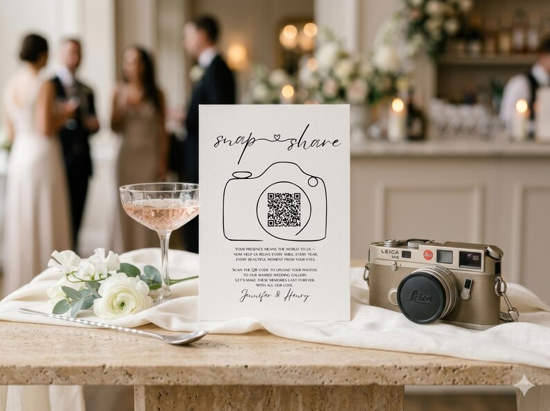 May include: A white rectangular wedding sign with black text reading snap share, a line-art camera icon with a QR code, and the names Jennifer & Henry. The sign is displayed next to a vintage gold Leica camera and a glass of pink liquid.
