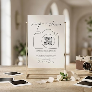 May include: A white rectangular sign in a gold metal stand featuring the text snap share, a line-art camera icon with a QR code in the center, and the names Jennifer & Henry. The sign includes instructions for guests to upload photos.