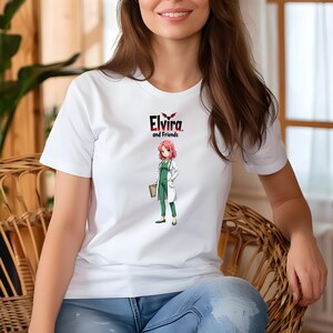 May include: White t-shirt featuring a graphic of an anime-style character in a green jumpsuit and white lab coat, holding a folder. The shirt has the text "Elvira and Friends" above the character. The design includes a bat graphic.