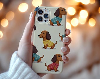 Peut inclure: Une coque de téléphone avec un motif répété de teckels de dessins animés portant des pulls de différentes couleurs. Les teckels ont une fourrure brune et des oreilles tombantes. La coque est tenue par une personne.