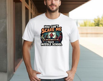 May include: White t-shirt with the text "YOU CAN'T SCARE ME I TEACH MIDDLE SCHOOL" in orange and black. The graphic depicts a teacher in front of a classroom. The shirt is worn by a person.