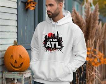 Puede incluir: Sudadera con capucha blanca con el texto "I'M SO ATL" en blanco y negro, con un sol rojo y una silueta de horizonte. La sudadera tiene una capucha con cordón y un bolsillo delantero. La persona lleva vaqueros azules.