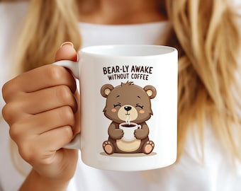 Op de afbeelding: Witte keramische mok met een cartoonbeer die een koffiekopje vasthoudt. De mok heeft de tekst "BEAR-LY AWAKE WITHOUT COFFEE". De beer is bruin met beige accenten.