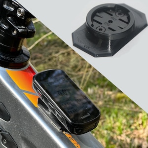Könnte beinhalten: Schwarzer Garmin Fahrradcomputer an einem Fahrradrahmen montiert, der Daten anzeigt. Eine separate schwarze Kunststoffhalterung ist ebenfalls zu sehen. Der Fahrradrahmen ist silberfarben mit orangefarbenen und gelben Akzenten. Der Bildschirm zeigt Zeit, Distanz und Geschwindigkeit.