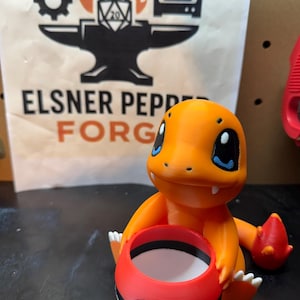 May include: An orange Charmander figure holding a red and white Poké Ball. The figure has black eyes and white claws. The background features a sign with the text "Elsner Pepper Forge" and a logo with a gear, anvil, and a 20-sided die.