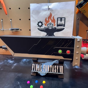 May include: A black and tan Buster Sword replica from Final Fantasy VII, with green and red button details. The sword rests on a stand with the game's logo and a few colorful spheres.