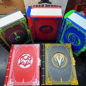 May include: A collection of colorful, decorative books with intricate designs. Each book features a unique color scheme, including red, blue, green, and gray, with detailed embellishments and symbols. The books are arranged on a dark surface.