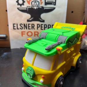 May include: A 3D-printed toy van in the style of the Teenage Mutant Ninja Turtles' vehicle, the Party Wagon. The van is primarily yellow with green and gray accents, featuring the Turtles logo on the front. The background includes a logo and the text "ELSNER PEPPE FOR".