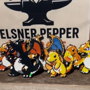 May include: Pixelated character keychains in various colors, including orange, yellow, black, and blue. The keychains are arranged in a row on a wooden surface. The background features a logo with the text "ELSNER PEPPER".