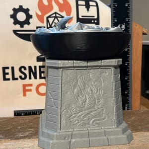 May include: A gray 3D-printed decorative bowl with a black rim, resting on a gray pedestal. The bowl contains a gray landscape with a small creature. The pedestal features an engraved design. The background includes a ruler and a logo.