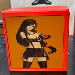 Op de afbeelding: Een felrode vierkante spaarpot met een plat, oranje paneel aan de voorzijde. Het paneel toont een gedetailleerde grafische illustratie van een langharig personage met een wit hemdje, een donkere rok en handschoenen, uitgevoerd in bruintinten.