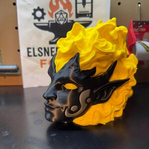 May include: A 3D-printed sculpture of a stylized face with yellow hair and black facial features. The face has pointed ears and a decorative design on the cheek. The sculpture is set against a backdrop with the text "ELSNE F".