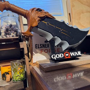 May include: A detailed replica of a God of War blade, featuring a brown handle and a black and gray blade with gold accents. The base displays the God of War logo. The text "ELSNER FORGE" is also visible.