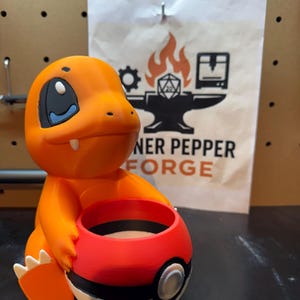 May include: An orange Charmander figure holding a red, black, and white Poke Ball. The figure has a blue eye and white claws. A paper bag in the background has a logo with flames and the text "NER PEPPER FORGE".