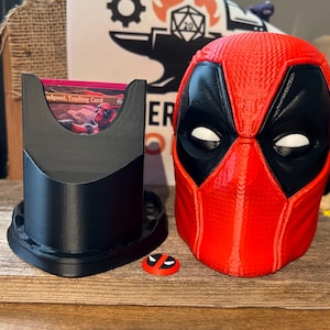 May include: A red and black Deadpool mask, a black card holder with a trading card, and a small Deadpool logo are displayed on a wooden surface. The mask is detailed with white eye shapes. The card holder has a curved design.