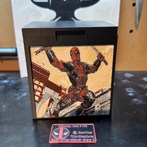 Può includere: Una scatola portaoggetti quadrata nera con un'illustrazione del personaggio Deadpool che impugna due spade sul pannello frontale. Davanti alla scatola si trova una piccola targa rettangolare rossa con il testo: Upkeep, lose 3 life, 0, Sacrifice This Creature: Each other player draw 1.