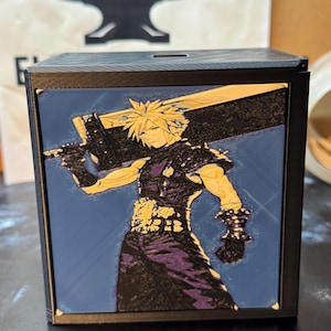 May include: A black square box featuring a multi-colored 3D printed relief of a spiky-haired character holding a large sword across their back. The character wears dark clothing with textured details, set against a flat blue background panel.