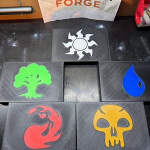May include: A collection of black rectangular plaques, each featuring a different colored symbol. The symbols include a white sun, a green tree, a blue water droplet, a red flame, and a yellow skull. The text "ELSNER PEPPER FORGE" is visible in the background.