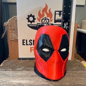 May include: A red and black Deadpool mask, with white eye cutouts, sits on a wooden surface. The mask is 3D-printed and has a textured surface. A ruler is placed behind the mask, and a sign with a logo is visible in the background.