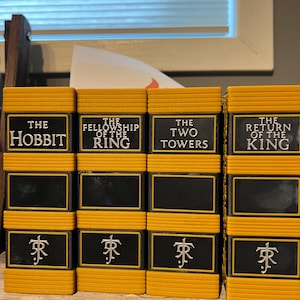 May include: A stack of four yellow and black bookends, each labeled with a title from the Lord of the Rings and The Hobbit series. The titles are in white text on a black background. The bottom of each bookend has a white symbol.