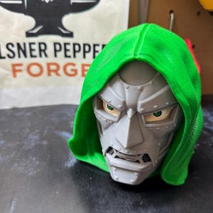 Op de afbeelding: Een 3D-geprint borstbeeld van een personage met een grijs, metaalkleurig masker met zichtbare klinknagels en een felgroene capuchon. Het personage heeft scherpe, hoekige gelaatstrekken, een prominente neus en zichtbare groene ogen.