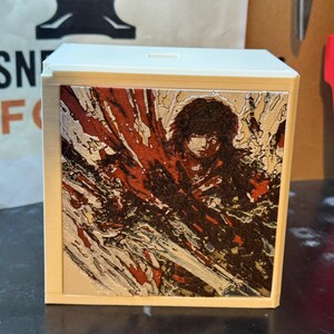 May include: A white square box featuring a recessed front panel with a detailed, multi-colored graphic print. The image depicts a dark-haired figure in a dynamic pose, surrounded by abstract, flowing patterns in shades of red, silver, and black.