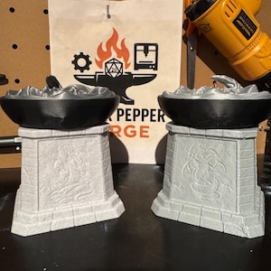May include: Two 3D-printed candle holders featuring a black bowl-shaped top resting on a light gray, stone-textured pedestal base. Each pedestal displays a relief carving of a dragon on the front face, with a smooth, matte finish on the bowl.