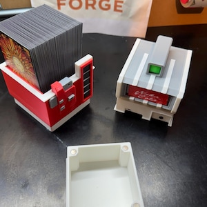 May include: A red and white card holder with a stack of cards, a gray and white card holder with "Nuka Cola" text, and a white box with magnets. The sign in the background says "Elsner Pepper Forge."