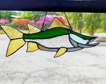 May include: A stained glass fish suncatcher featuring a green back, a textured white belly, and yellow fins and tail. The piece is constructed with dark metal solder lines and includes a black metal chain for hanging, showcasing a multi-colored design.