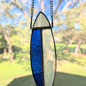 Peut inclure: Pendentif en forme de planche de surf en vitrail avec une chaîne argentée. Le pendentif est divisé en sections de verre blanc et bleu. La section supérieure est un triangle blanc. La forme de la planche de surf mesure environ 15 cm de long.