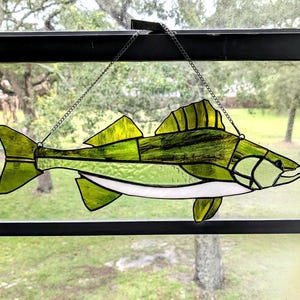 May include: A stained glass suncatcher shaped like a fish, featuring various shades of green glass with a white underbelly and dark solder lines. The piece is suspended by a thin metal chain, showcasing a textured finish and translucent quality.