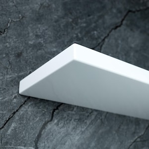 Könnte beinhalten: Ein weißes, rechteckiges Regal mit sauberem, modernem Design. Das Regal ist an einer dunkelgrauen, strukturierten Wand befestigt. Die Abmessungen des Regals betragen ca. 30 cm Länge und 15 cm Breite.
