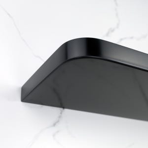 Può includere: Primo piano di una mensola nera curva con finitura liscia e lucida. La mensola ha angoli arrotondati ed è montata su una superficie bianca con motivo marmoreo. Il design della mensola è minimalista e moderno.