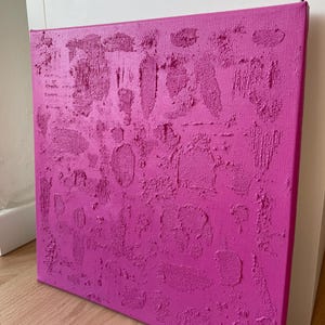 Puede incluir: Una obra de arte en lienzo cuadrada de color magenta con una superficie texturizada. La textura en relieve crea un patrón irregular de formas y líneas. El lienzo está estirado sobre un bastidor de madera.