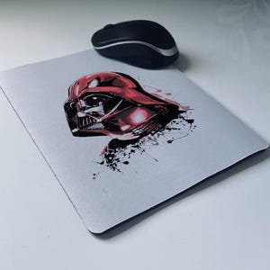 Könnte beinhalten: Rechteckiges Mauspad mit einer Grafik von Darth Vaders Helm in Rot und Schwarz. Eine schwarze Computermaus liegt darüber. Das Mauspad hat einen schwarzen Rand.