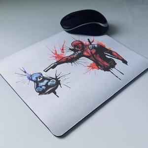 May include: White rectangular mousepad featuring a Deadpool graphic in red and black, with a blue cartoon character. A black wireless mouse sits above the mousepad. The mousepad has a black border.