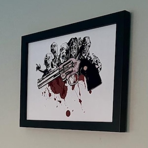 May include: Framed artwork depicting a black and white illustration of zombies and a revolver with blood splatter. The artwork is in a black frame and is mounted on a wall.