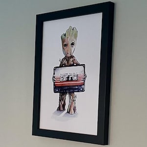 May include: Framed artwork featuring a watercolor illustration of Groot holding a retro cassette tape. The tape is labeled "Awesome Mix Vol. 2" in white text. The artwork is set in a black frame.
