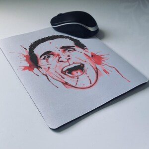 May include: A white mousepad featuring a graphic illustration of a screaming face with red blood splatter details. A black wireless computer mouse is positioned above the mousepad.